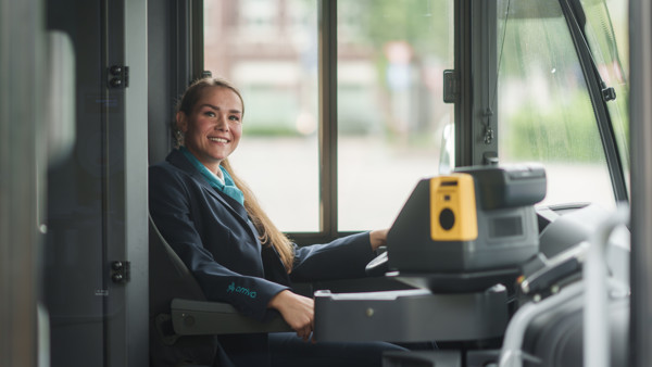 Buschauffeur zit achter stuur van een bus en kijkt lachend opzij in de camera. Buschauffeur zit achter stuur van een bus en kijkt lachend opzij in de camera.